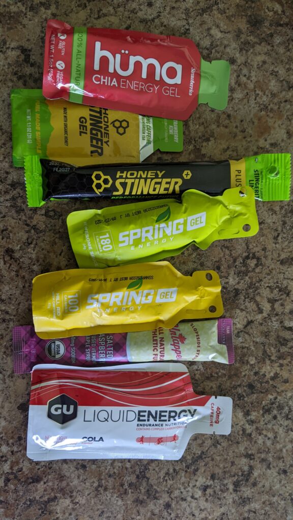 A picture of six energy gels and one pack of energy chews. The gels are Huma Strawberry, Honey Stinger Strawberry-Kiwi, Spring Awesome Sauce, Spring "Hill Aid" Mango, Untapped Maple Salted Raspberry, and Gu Cola. The chews are Honey Stinger "Stingerita Lime"